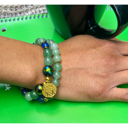 Lifestyle photo of a hand wearing a double-stacked Gemini zodiac bracelet set made with green aventurine and dyed blue agate beads, featuring a gold Gemini charm, photographed on a green notebook background.